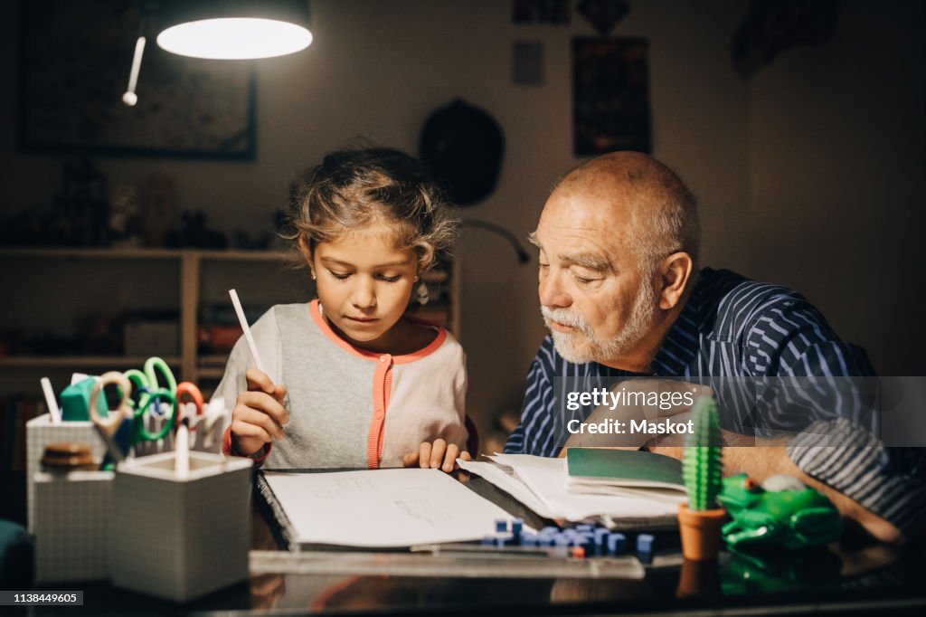Grandfather looking at granddaughter writing homework on desk in house