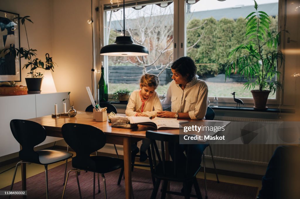 Mother assisting daughter in writing homework while sitting at home