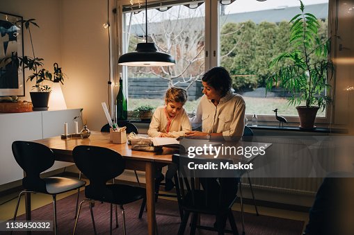 Mother assisting daughter in writing homework while sitting at home Mother assisting daughter in writing homework while sitting at home