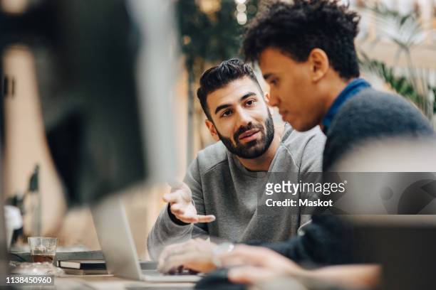 male computer programmers discussing over laptop on desk while sitting in office - collega d'ufficio foto e immagini stock
