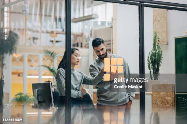 businessman discussing with businesswoman sticking adhesive notes on glass wall in office - entwicklung stock-fotos und bilder