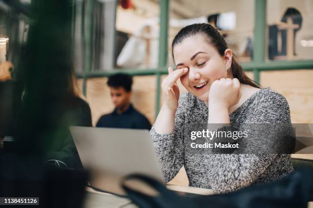 smiling businesswoman rubbing eyes while using laptop on desk in office - rubbing eyes stock pictures, royalty-free photos & images