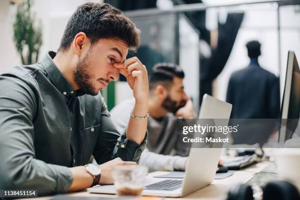 exhausted businessman looking at laptop on desk while sitting with male coworker in office - agotamiento fotografías e imágenes de stock
