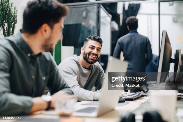 smiling male colleagues looking at each other while sitting by desk in office - working behind laptop fotografías e imágenes de stock