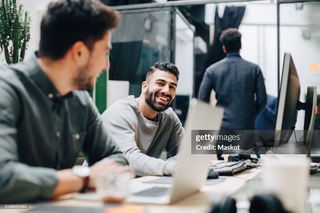 Smiling male colleagues looking at each other while sitting by desk in office
