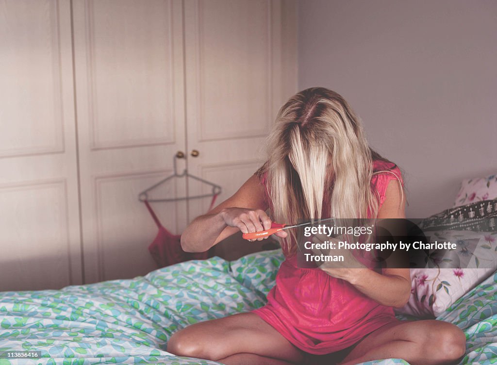 Blonde woman sitting on bed cutting her hair