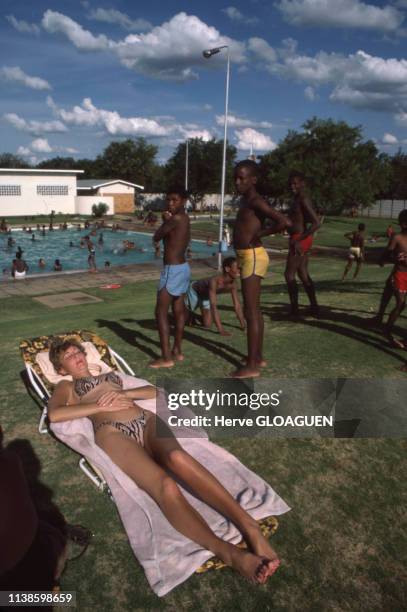 Jeune femme blanche bronzant allongée sur une serviette au bord d'une piscine pendant que des enfants noirs jouent sur la pelouse près d'elle durant...