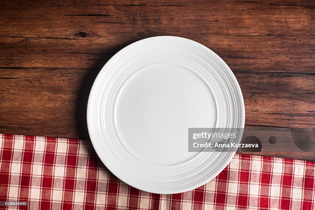 Empty white plate on a napkin on an old wooden brown background, top view. Image with copy space. Kitchen table with a towel and a plate - top view with copy space.