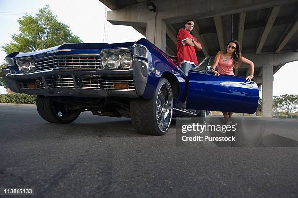 portrait of a young hip-hop couple standing beside a pimped-up vintage car under a highway overpass - pimped car foto e immagini stock