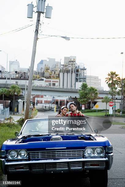 portrait of a young hip-hop couple sitting in a pimped-up vintage car in depressed urban neighborhood - pimped car foto e immagini stock