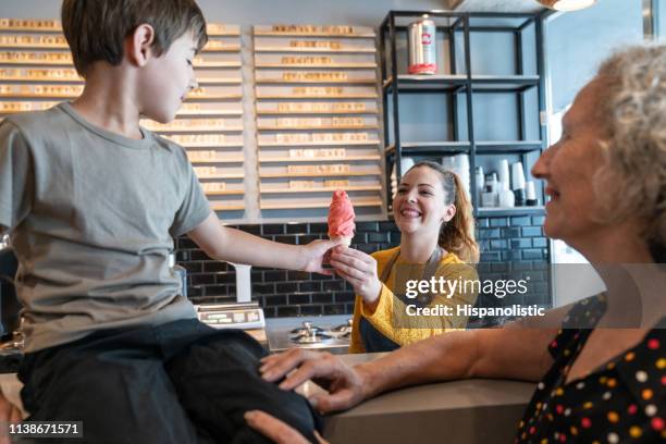 freundliche mitarbeiterin überreicht einen eiskegel an den jungen, der auf der theke sitzt, und großmutter neben ihm alle lächelnd - ice cream counter stock-fotos und bilder