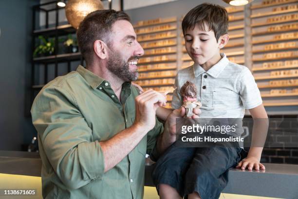 fröhlicher papa füttert seinen sohn eis, während er auf theke in der eisdiele sitzt - ice cream counter stock-fotos und bilder