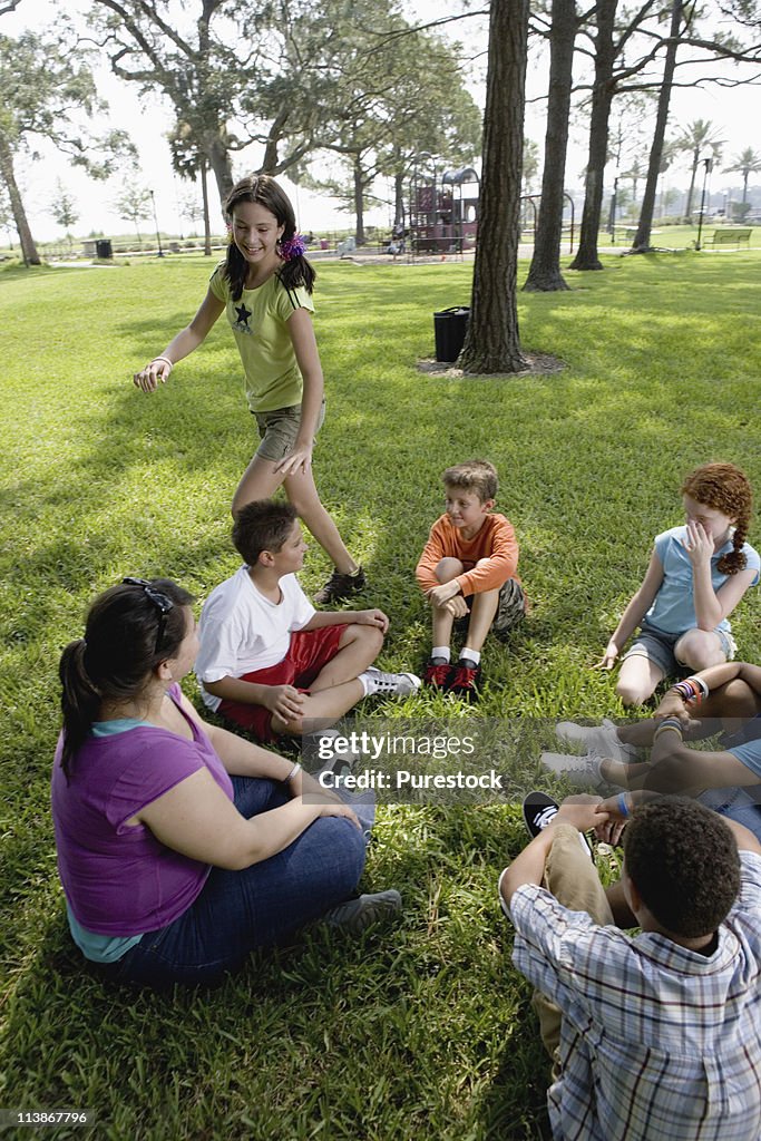 Children sitting in a circle on the grass with their teacher while a girl walks around them