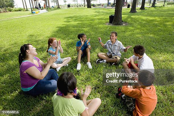 children sitting in a circle on the grass with their teacher at a park - enfant chant classe photos et images de collection