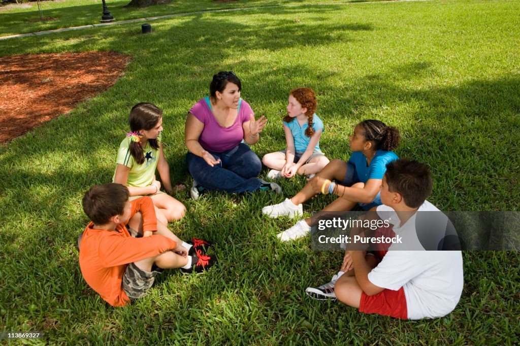 Children sitting in a circle on the grass with their teacher at a park