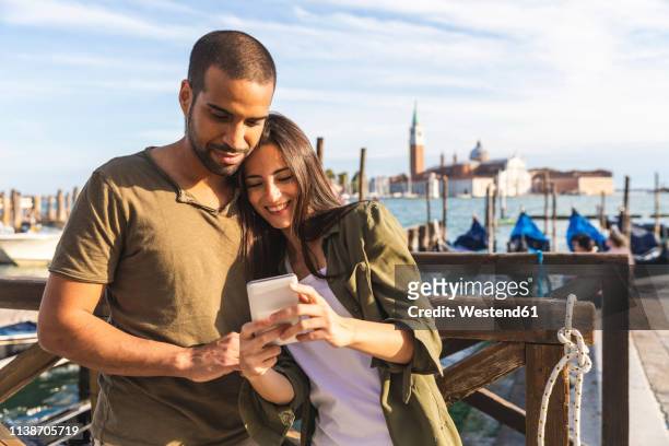 italy, venice, affectionate young couple with cell phone and gondola boats in background - passagier wasserfahrzeug stock-fotos und bilder