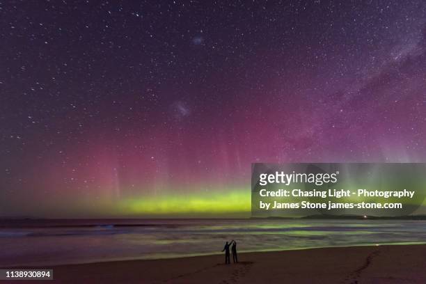 a couple in silhouette dancing on a beach watching an incredible bright green display of the aurora australis or southern lights over a beach in tasmania with bright blue bioluminescence in the waves. - aurora australis stock pictures, royalty-free photos & images