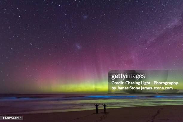 a couple in silhouette standing on a beach watching an incredible bright green display of the aurora australis or southern lights over a beach in tasmania with bright blue bioluminescence in the waves. - southern lights stock pictures, royalty-free photos & images