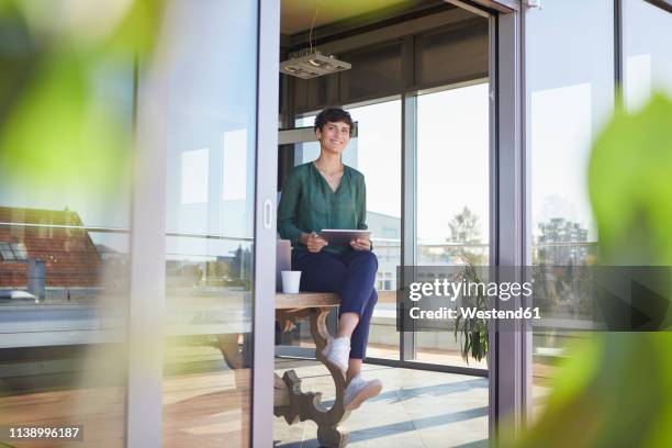 smiling businesswoman sitting on table with tablet - sunny office stock pictures, royalty-free photos & images