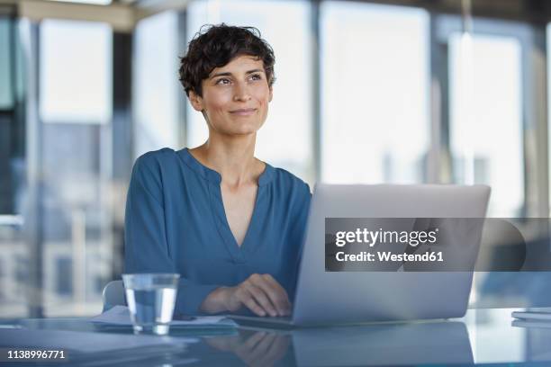 smiling businesswoman sitting at desk in office with laptop - looks right stockfoto's en -beelden