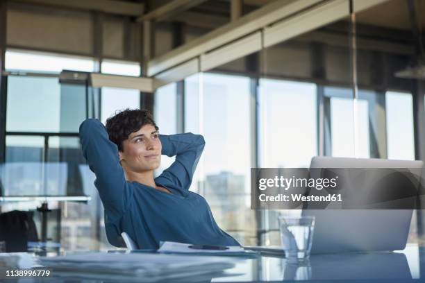 smiling businesswoman sitting at desk in office with laptop having a break - hands behind head stock pictures, royalty-free photos & images