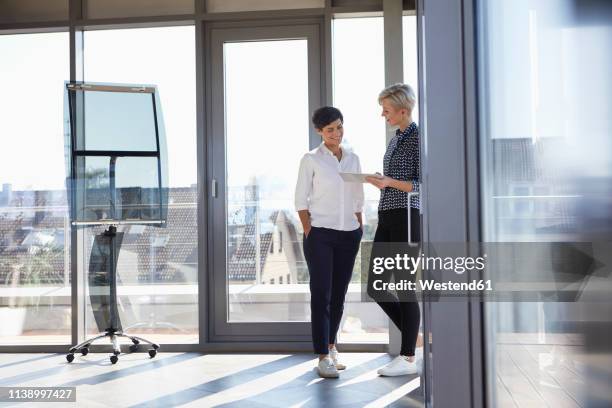 two smiling businesswomen looking at tablet at the window in bright office - man standing talking stock-fotos und bilder