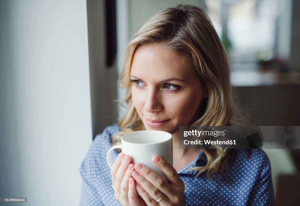 Smiling woman holding a cup of coffee at the window at home