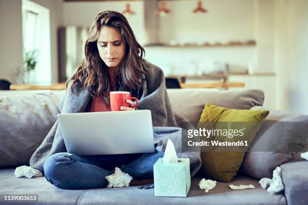 sick woman sitting on sofa covered in blanket with cup of tea and laptop - krankheit stock-fotos und bilder