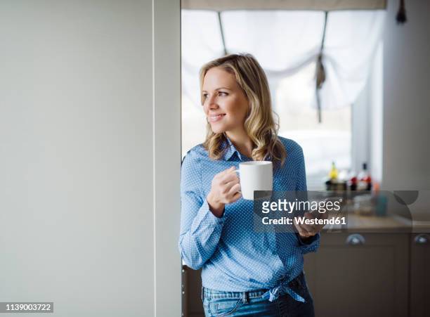 smiling woman holding a cup of coffee and smartphone at home - woman drinking phone kitchen stockfoto's en -beelden