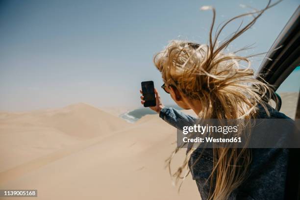 namibia, walvis bay, namib-naukluft national park, sandwich harbour, woman leaning out of car window taking cell phone picture - one man only photos stock-fotos und bilder