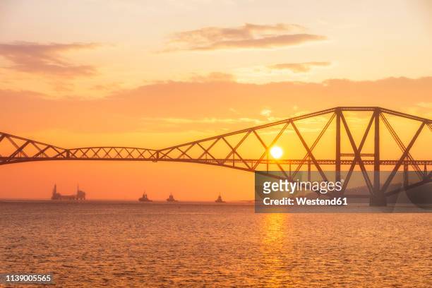 united kingdom, scotland, firth of forth, forth rail bridge with tug boats underneath and hound point oil loading marine terminal at sunset - south queensferry ストックフォトと画像