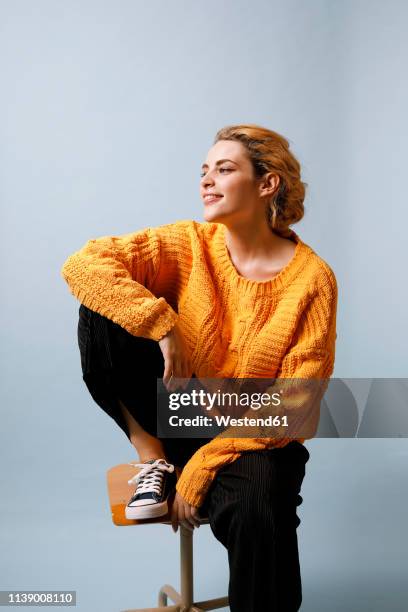 smiling young woman sitting on chair in front of blue background wearing yellow knit pullover - sedersi posizione fisica foto e immagini stock