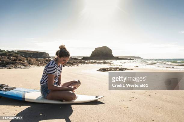 young woman on the beach, sitting on surfboard, using smartphone - woman smartphone nature stock pictures, royalty-free photos & images