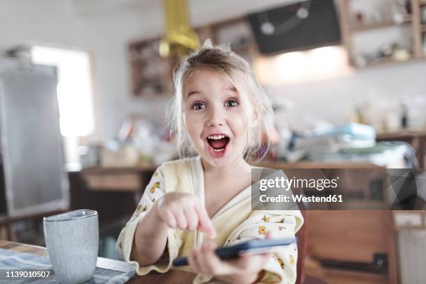 portrait of excited little girl in the kitchen pointing at smartphone - very young girls stock-fotos und bilder