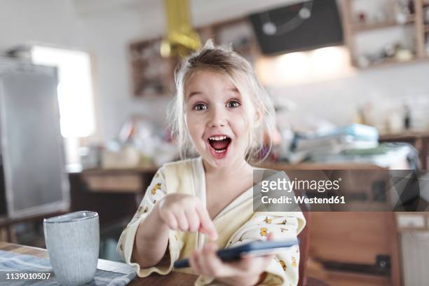 portrait of excited little girl in the kitchen pointing at smartphone - mobile bad news stock pictures, royalty-free photos & images