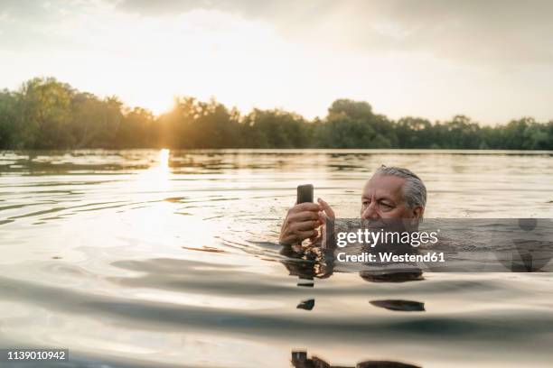 senior man swimming in a lake at sunset using cell phone - senior swimming stock pictures, royalty-free photos & images