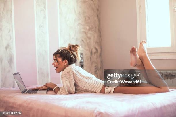 surprised young woman lying on bed using laptop - allongé sur le devant photos et images de collection