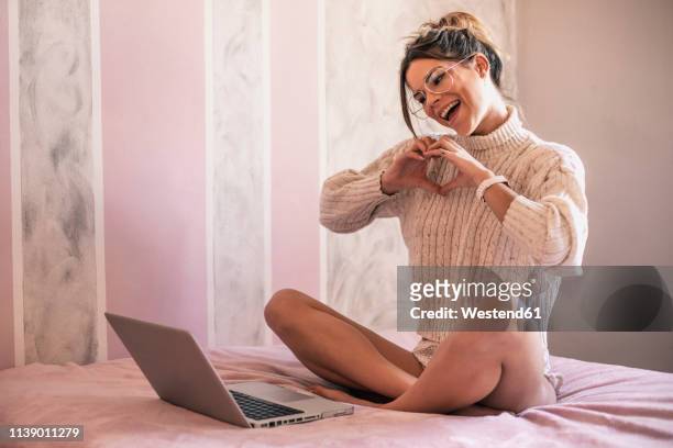 young woman sitting on bed with laptop forming a heart with her hands during video chat - women in slips stock-fotos und bilder