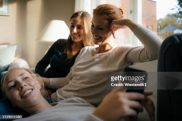 happy mother with two teenage girls on couch at home with cell phone - sister day stock pictures, royalty-free photos & images