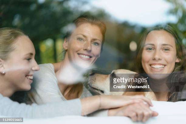 portrait of happy mother with two teenage girls and dog behind windowpane - mutter kind lachen zuhause stock-fotos und bilder