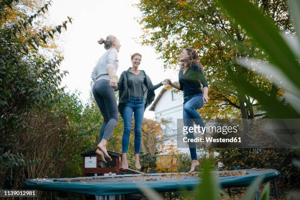 happy mother with two teenage girls jumping on trampoline in garden in autumn - trampoline equipment stock-fotos und bilder