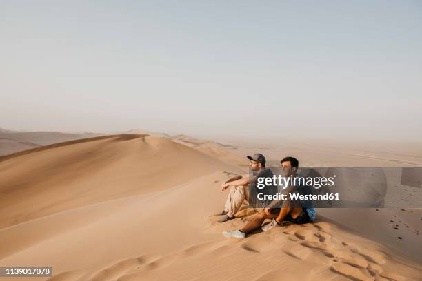 namibia, namib, two friends sitting on desert dune looking at view - walvis bay stock pictures, royalty-free photos & images