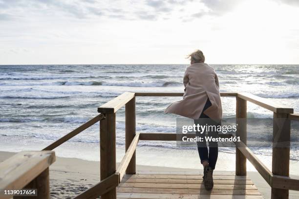 spain, menorca, back view of senior woman standing on boardwalk in winter looking at the sea - boardwalk stock-fotos und bilder