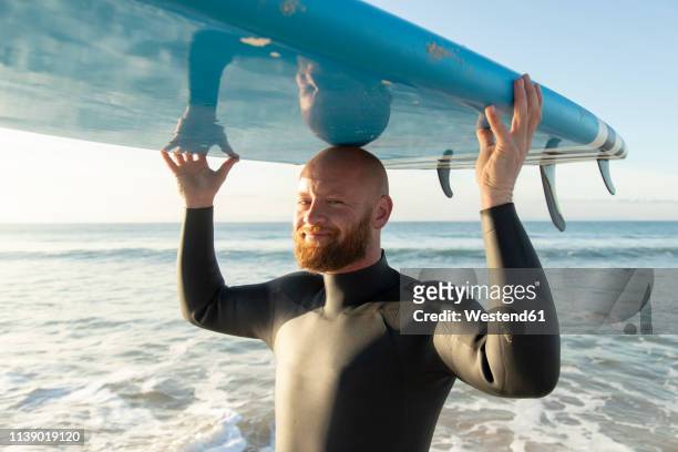 spain, andalusia, tarifa, portrait of smiling man carrying stand up paddle board at the sea - bald 30s stockfoto's en -beelden