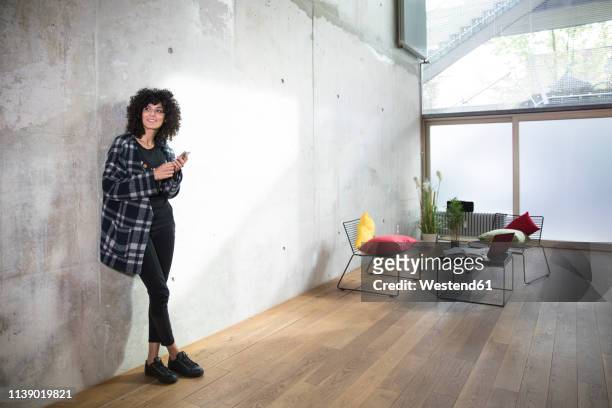 smiling woman with cell phone leaning against concrete wall in a loft - phone leaning stock pictures, royalty-free photos & images