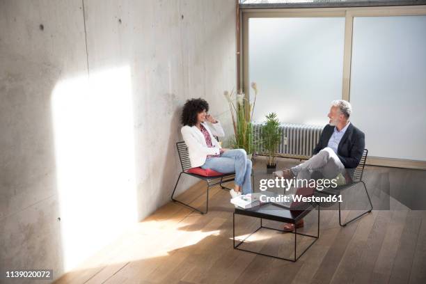 businessman and businesswoman sitting in a loft at concrete wall talking - sitting chair office relax stockfoto's en -beelden