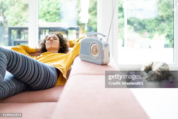 woman lying on couch listening to music with portable radio at home - poste de radio photos et images de collection