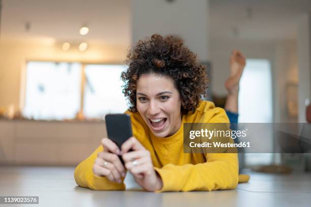 excited woman lying on the floor at home using cell phone - ropa tirada en el suelo fotografías e imágenes de stock