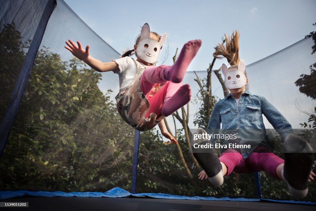 Two children wearing rabbit masks and bouncing on a trampoline