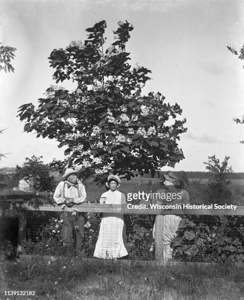 Outdoor portrait of the August Krueger family posing with farming tools and leaning on a fence in front of a Catalpa tree, Emmet, Wisconsin, 1908....